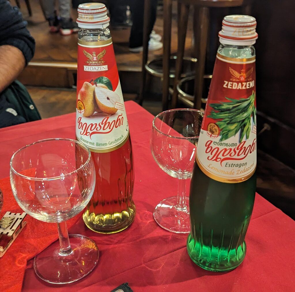 Zwei georgische Limonaden stehen samt Gläsern auf einem Tisch mit einer roten Tischdecke. Die Geschmackrichtungen sind Estragon und Birne.
